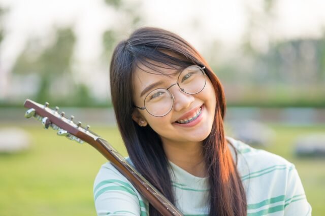 Asian teen girls with a guitar at the shoulder in the lawn. She is wearing braces and wear eyeglasses. How Does Wearing Braces Affect Playing an Instrument